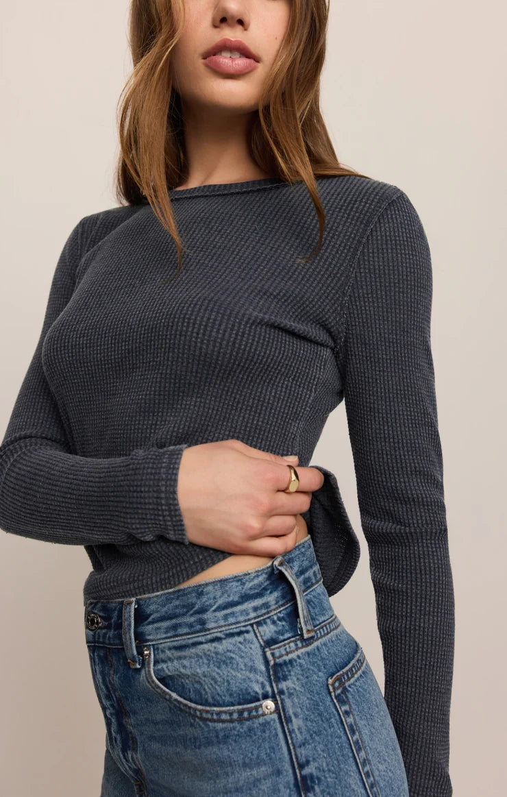 A woman wearing a dark charcoal gray ribbed long-sleeve crop top and blue jeans, cropped from the mouth down to the hips against a plain light beige background.