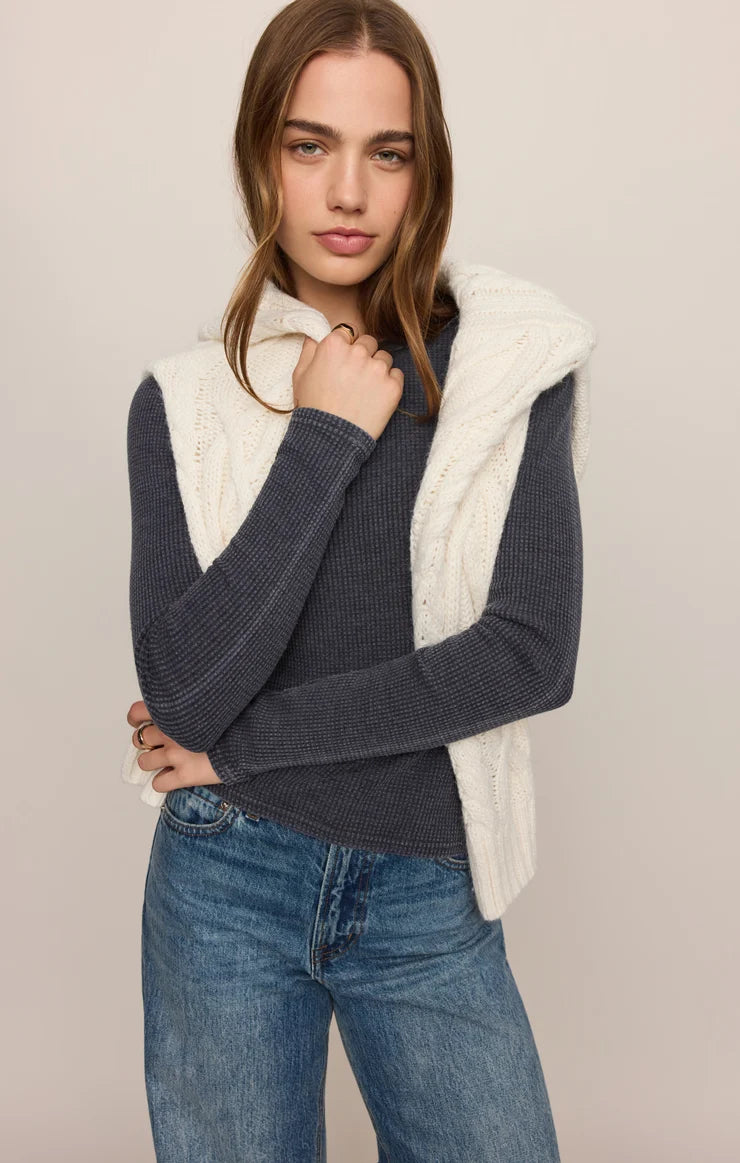 A woman modeling a navy blue ribbed knit sweater vest layered over a cream cable-knit hooded sweater, paired with blue jeans, against a solid light beige background.