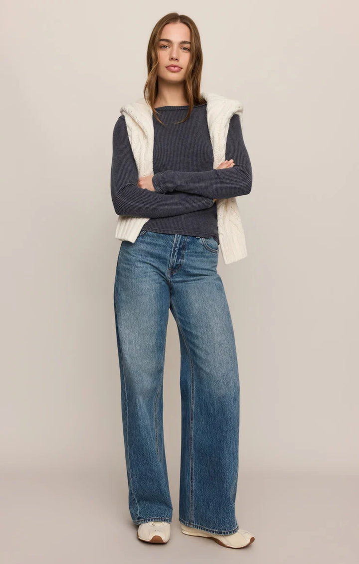 A woman stands against a plain light beige studio background wearing a dark navy long-sleeve top, a cream ribbed knit sleeveless vest, blue wide-leg jeans, and cream slip-on shoes.