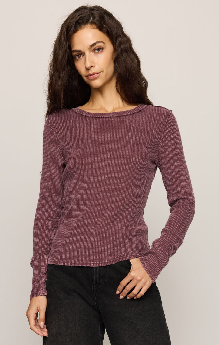 A woman modeling a long-sleeve ribbed knit top in a muted mauve-purple color, paired with black pants, against a plain light gray studio background.
