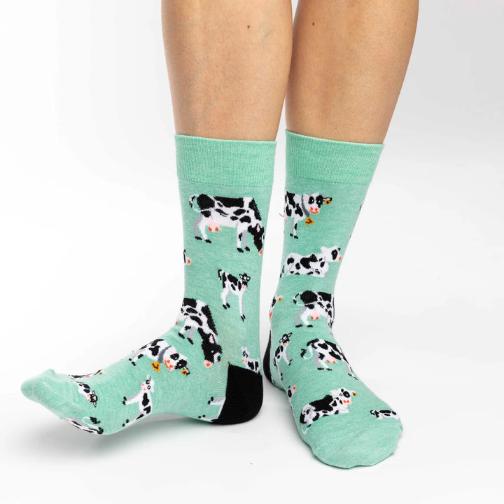 A pair of mint green crew socks with a black and white cow pattern, shown on a person&