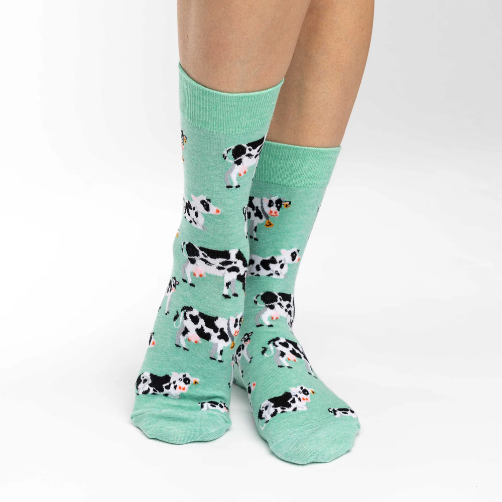 A pair of mint green crew socks with a black and white cow pattern, worn on a person's feet against a plain white background.