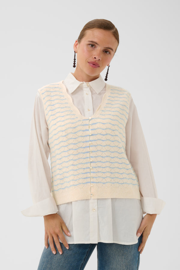 A woman modeling a cream-colored knit vest with light blue wavy horizontal stripes over a white collared shirt and blue jeans, set against a plain light gray studio background.