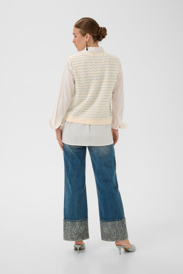 A woman is shown from the back wearing a cream knit vest with thin blue stripes over a white blouse, paired with wide-leg blue jeans featuring patterned cuffs, and silver heels, against a light gray studio background.