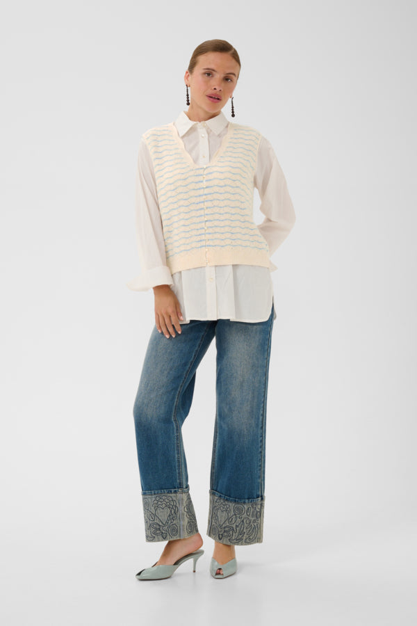 A model stands against a light gray studio background wearing a cream knit striped sweater vest over a white collared blouse, blue wide-leg jeans with patterned cuff detailing, and pale green pointed-toe heels.