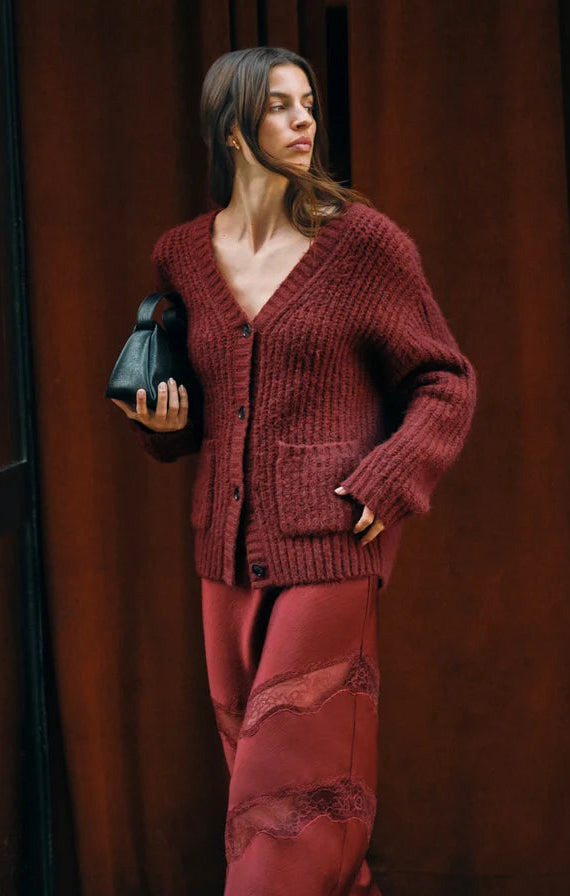 A woman stands outdoors wearing a chunky knit cardigan and matching satin-like skirt in a deep burgundy red, paired with black pointed ankle boots and holding a small black handbag.