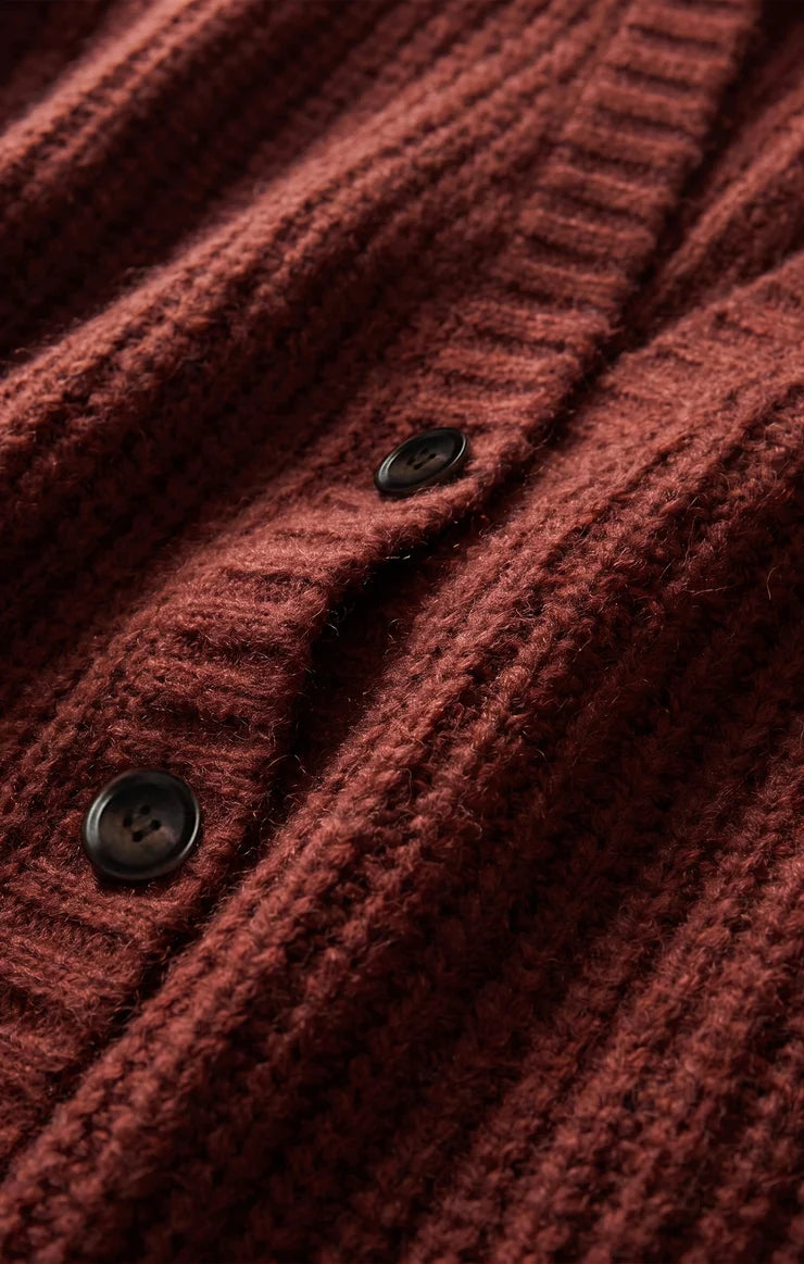 Close-up of a ribbed knit cardigan in a deep rust red color, featuring dark buttons and a soft, textured wool-like fabric.