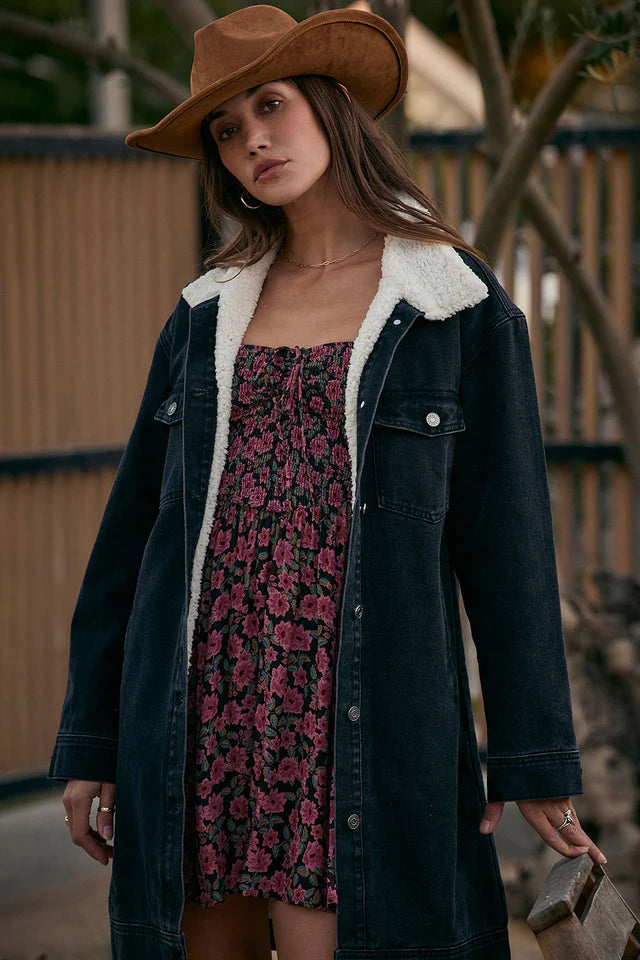 Woman wearing a floral dress, dark denim coat with white fur lining, and brown cowboy hat.