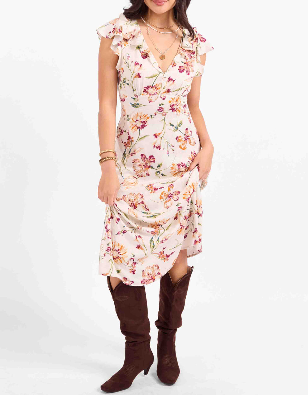 A woman models a sleeveless V-neck midi dress with a ruffled collar and a multicolor floral print on a light pinkish-beige background. She is wearing dark brown knee-high boots against a plain white studio background.
