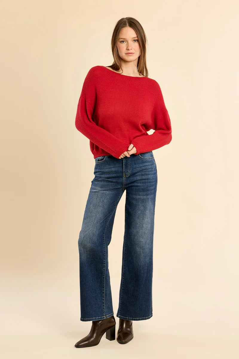 A woman modeling a bright red knit sweater with wide boat-style sleeves, paired with blue wide-leg jeans and dark brown heeled boots, against a solid beige studio background.
