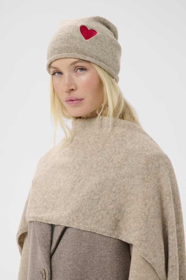 A woman wearing a beige knit beanie with a red heart patch and a matching beige wrap poncho over a taupe coat, photographed against a light gray studio background.