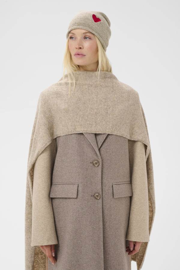 A model wearing a beige knit beanie with a small red heart, a taupe wool cape, and a matching taupe button-front coat, photographed against a light gray studio background.
