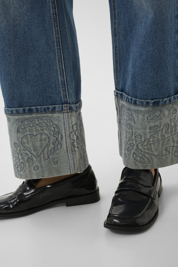 Close-up of jeans with embroidered cuffs and black loafers on a white background