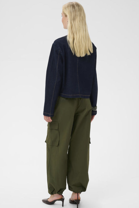 A woman with blonde hair is shown from the back wearing a dark navy cropped jacket, olive green cargo pants, and black heels against a light gray studio background.