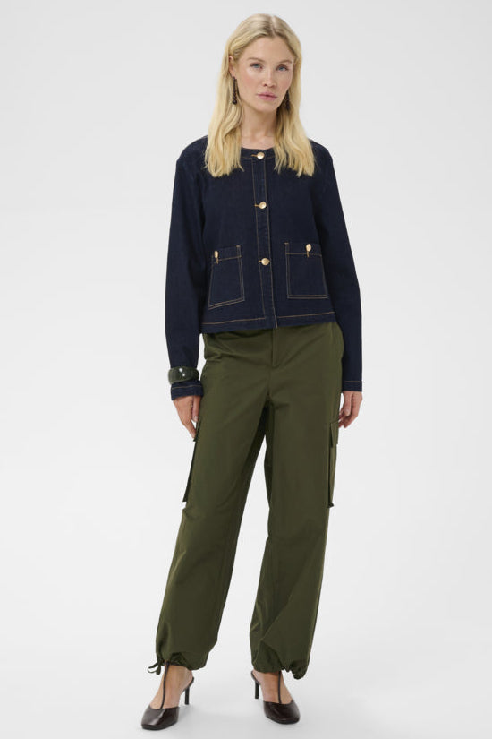 A blonde model stands against a light gray studio background wearing a dark navy cropped jacket with gold-tone buttons and contrast stitching, olive green cargo pants, and dark brown pointed-toe heels.