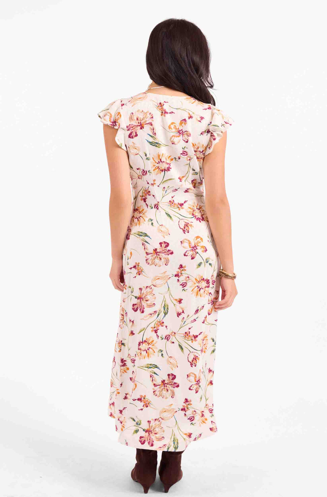 A woman viewed from the back wearing a sleeveless midi dress with ruffled cap sleeves and a floral print on a light cream background. The dress features pink, red, and green flowers with brown leaves.