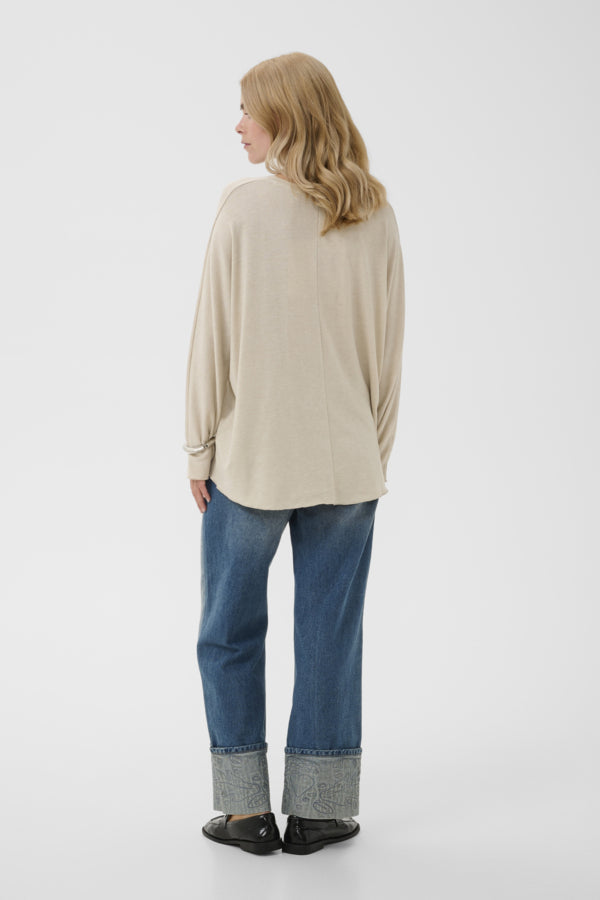 A woman shown from the back wearing a beige long-sleeve top, blue jeans with rolled cuffs, and black shoes, photographed against a plain light gray studio background.