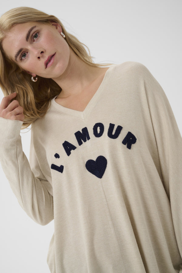 A woman wearing a beige long-sleeve V-neck sweater with 'L'AMOUR' and a heart printed in navy blue on the front, photographed against a light gray studio background.