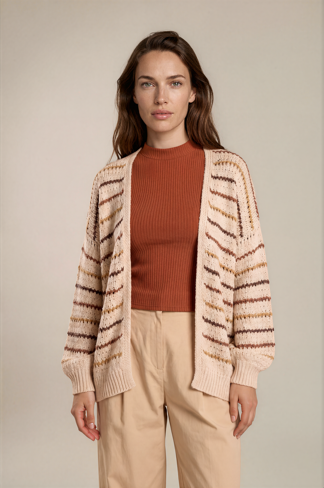 A woman stands against a plain beige studio background wearing a cream knit cardigan with brown and tan horizontal stripes over a rust-orange ribbed top and light beige pants.