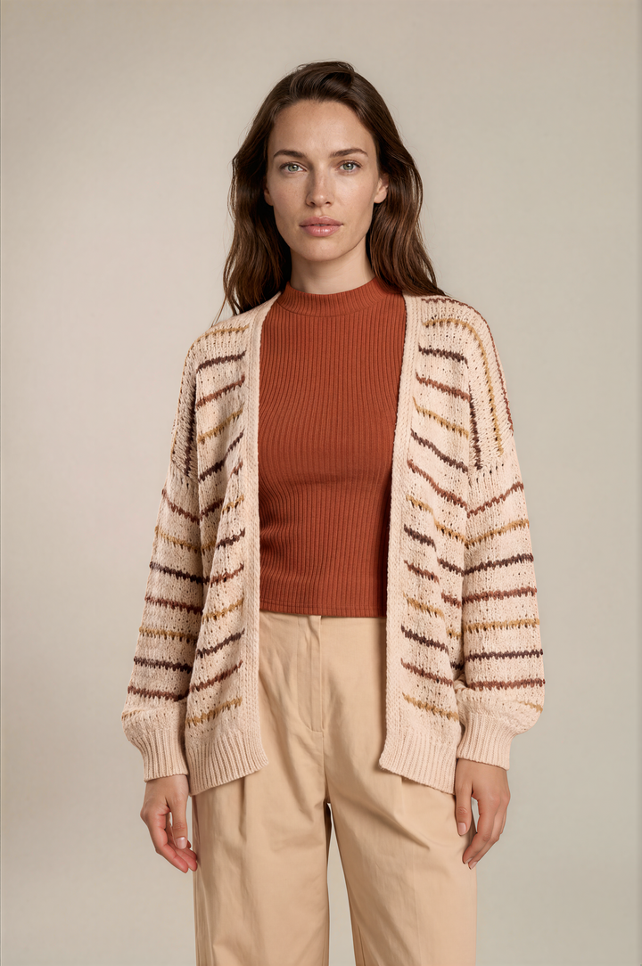 A woman stands against a plain beige studio background wearing a cream knit cardigan with brown and tan horizontal stripes over a rust-orange ribbed top and light beige pants.