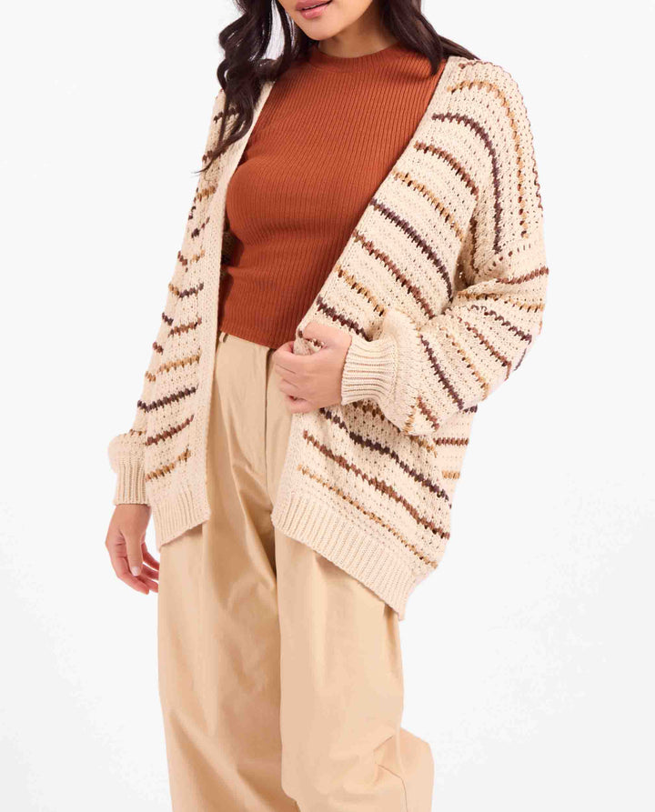 A woman wearing a cream knit cardigan with brown and beige horizontal stripes over a rust-orange ribbed top and beige pants, photographed against a plain white studio background.