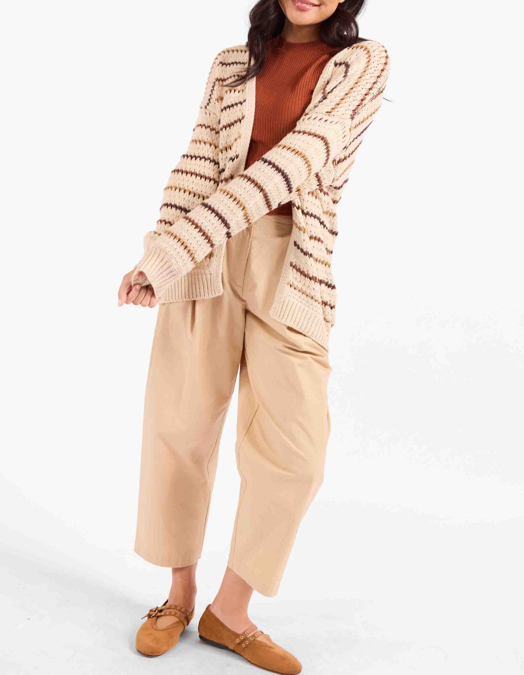 A woman modeling a beige and brown striped knit cardigan over a rust-brown top, paired with beige wide-leg cropped pants and tan loafers, against a plain white studio background.
