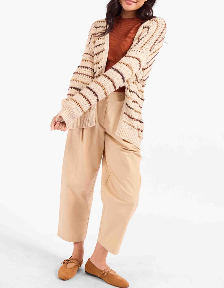 A woman modeling a beige and brown striped knit cardigan over a rust-brown top, paired with beige wide-leg cropped pants and tan loafers, against a plain white studio background.