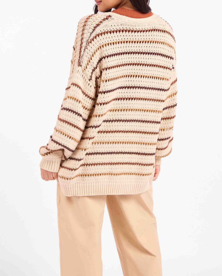A person shown from the back wearing an oversized chunky knit sweater with horizontal beige, cream, and brown stripes, paired with light beige pants, against a plain white background.