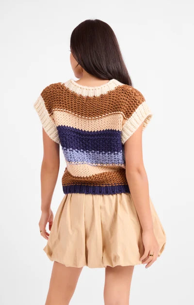 A woman shown from the back wearing a chunky knit striped sweater with brown, cream, and navy blue horizontal bands, paired with a pleated beige skirt, against a plain white background.