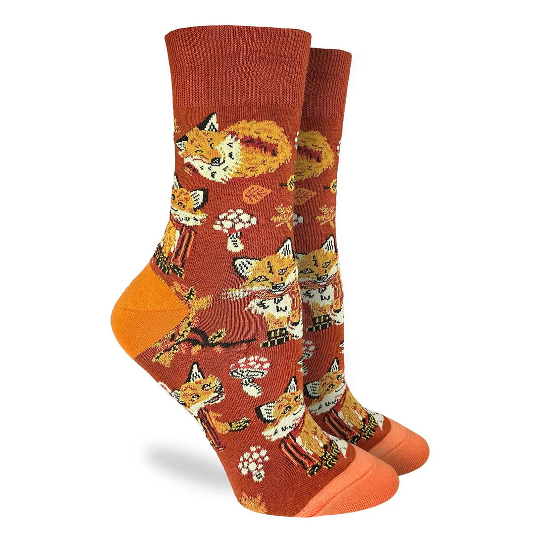 A pair of crew socks with a rust-orange background featuring a playful pattern of cartoon foxes, mushrooms, and autumn leaves, finished with solid orange toes and heels.
