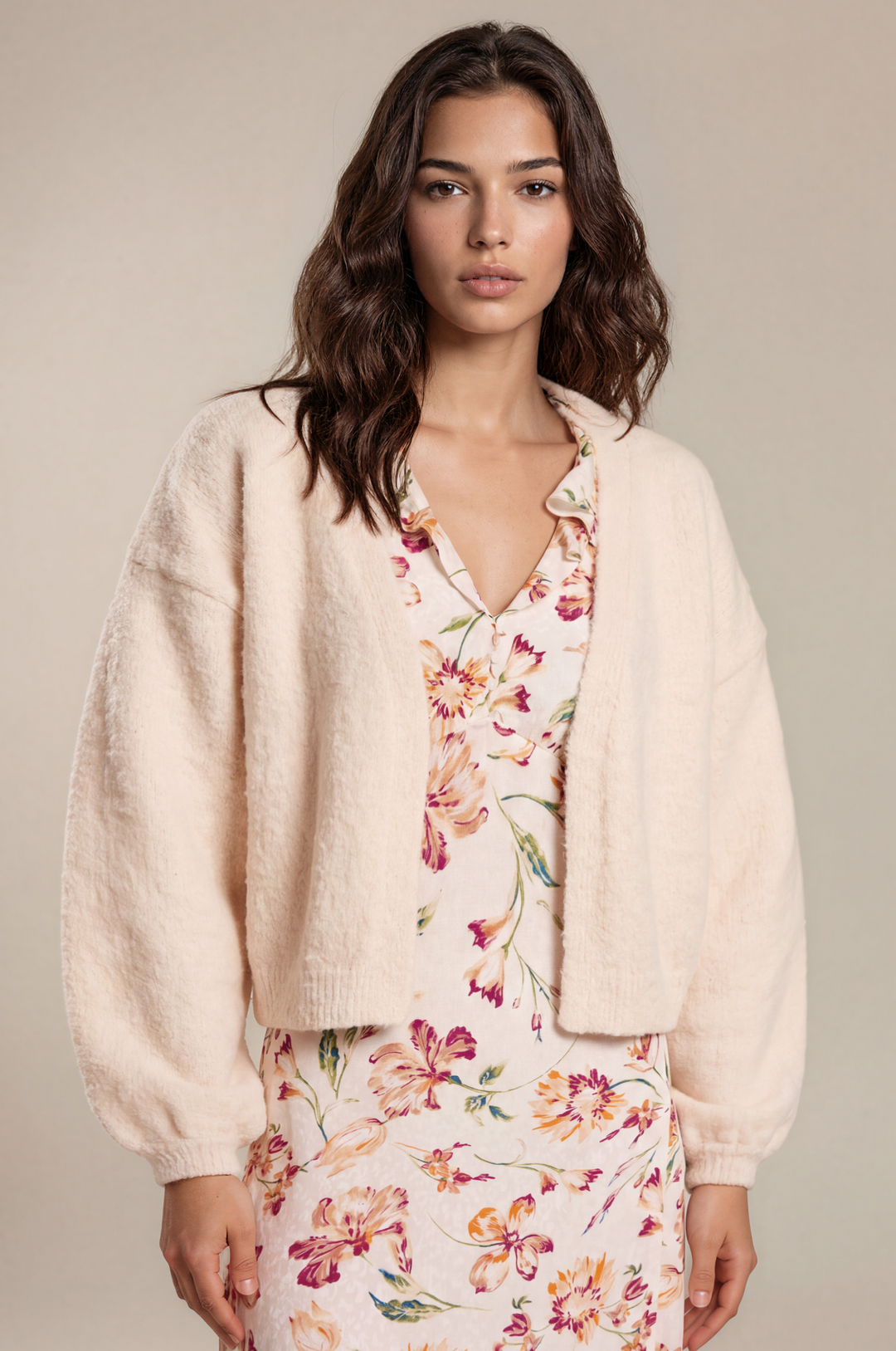 A woman modeling a cream-colored, fuzzy cropped cardigan over a matching cream floral dress, set against a solid light beige studio background.