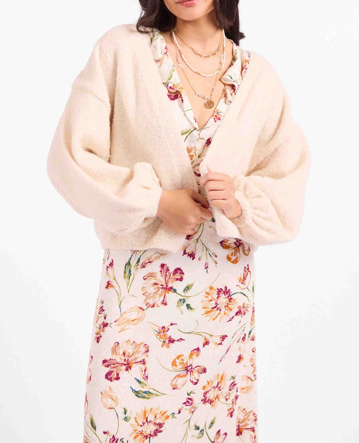 A woman wearing a cream fuzzy cropped cardigan over a floral print dress, photographed against a plain white background.