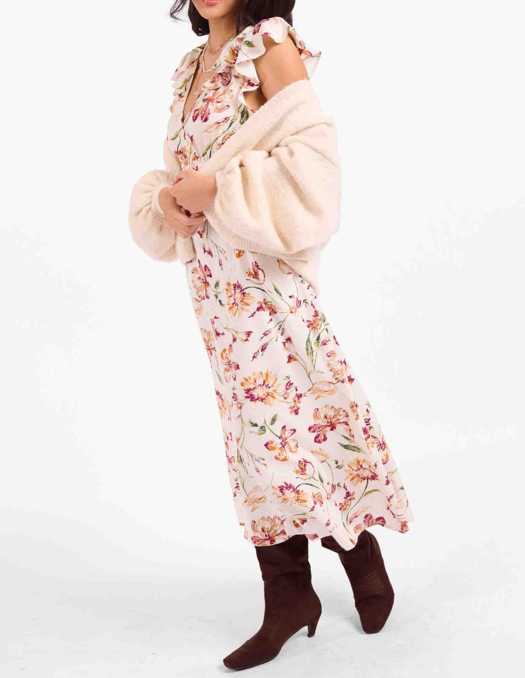 A woman models a long cream floral dress with ruffled shoulders and a matching fuzzy cream cardigan, paired with dark brown heeled boots, against a plain white studio background.