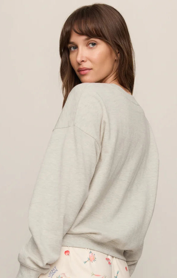 A woman wearing a light gray heather crewneck sweatshirt and cream shorts with a colorful fruit print, photographed against a solid light beige background.
