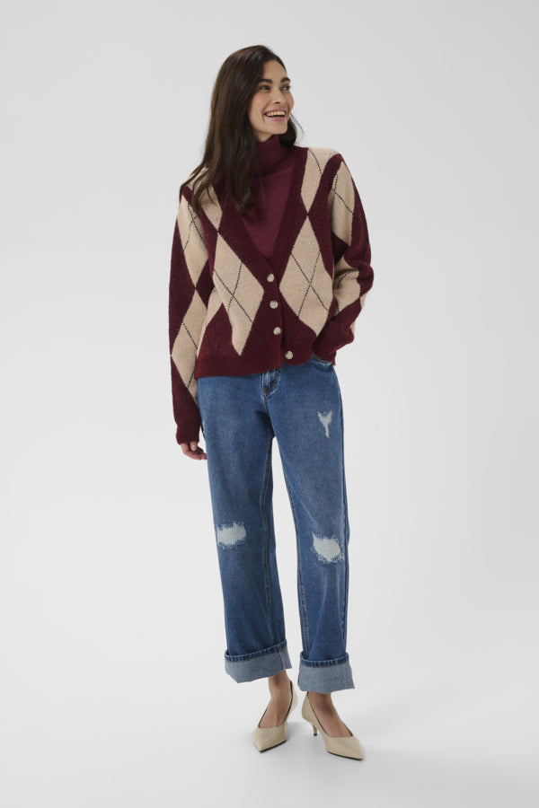 A woman modeling a burgundy and beige argyle cardigan over a matching burgundy turtleneck, paired with distressed blue jeans and beige heels, against a light gray studio background.