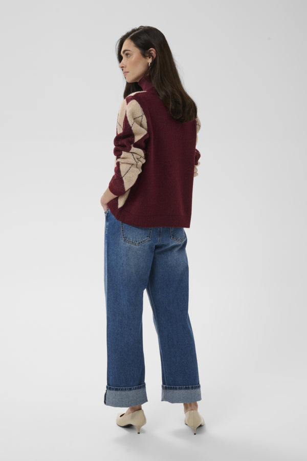 A woman is shown from the back wearing a burgundy knit sweater with beige geometric patterned sleeves, blue wide-leg jeans, and beige heels, against a light gray studio background.