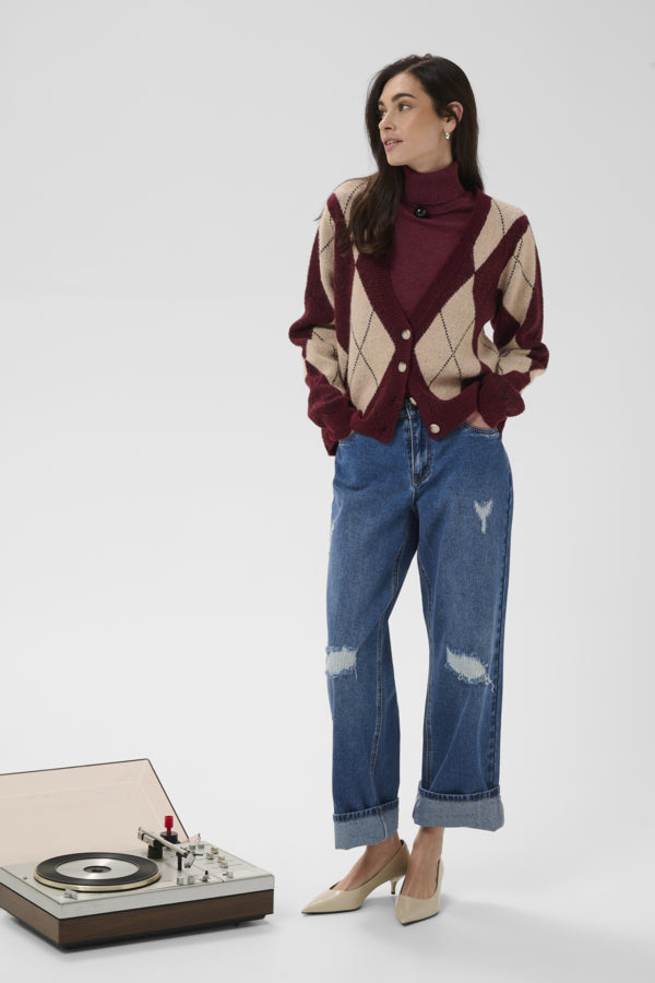 A woman stands in a studio wearing a burgundy and beige argyle cardigan over a burgundy turtleneck, blue distressed jeans, and beige heels. A turntable sits on the floor to the lower left against a light gray background.