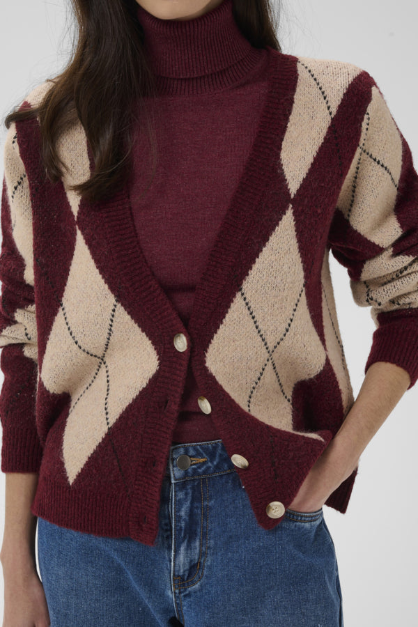 A close-up of a person wearing a burgundy and beige argyle cardigan over a burgundy turtleneck sweater, paired with blue jeans against a plain light gray background.