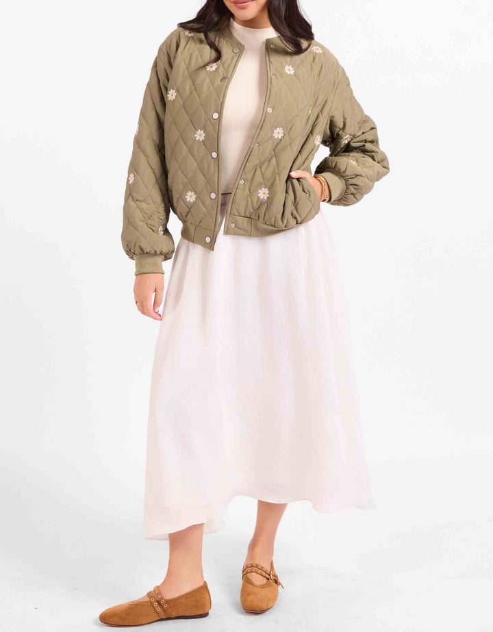 A model wearing a light olive green quilted bomber jacket with white floral embroidery, styled over a cream top and a light pink midi skirt, paired with brown suede flats, against a plain white background.