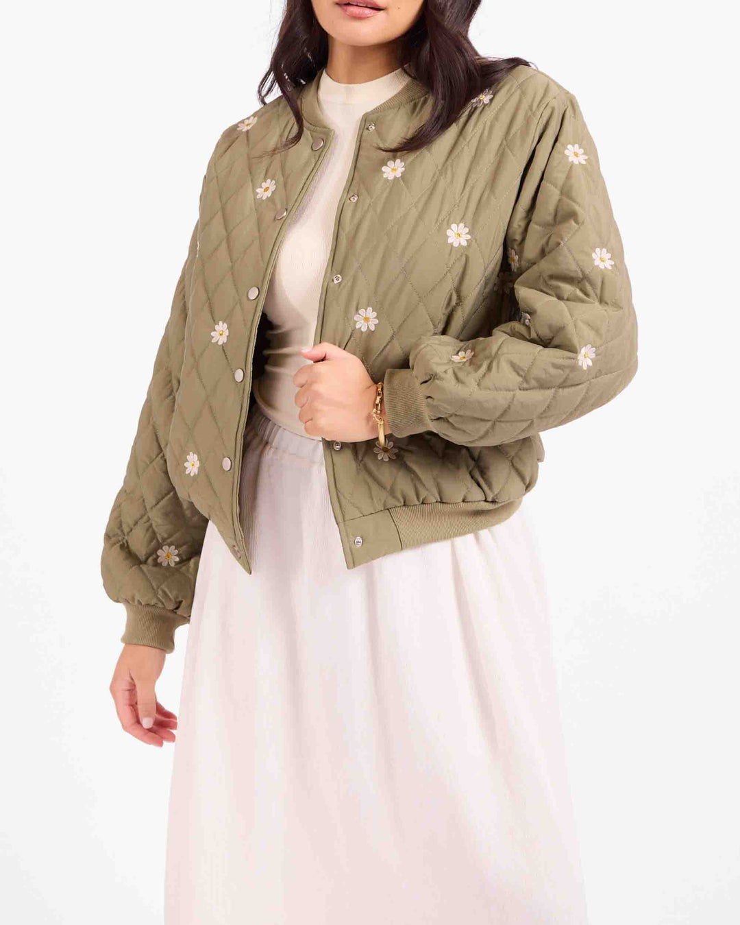 A woman wearing an olive green quilted bomber jacket with white embroidered flowers, styled over a cream top and a light pink skirt against a plain white background.