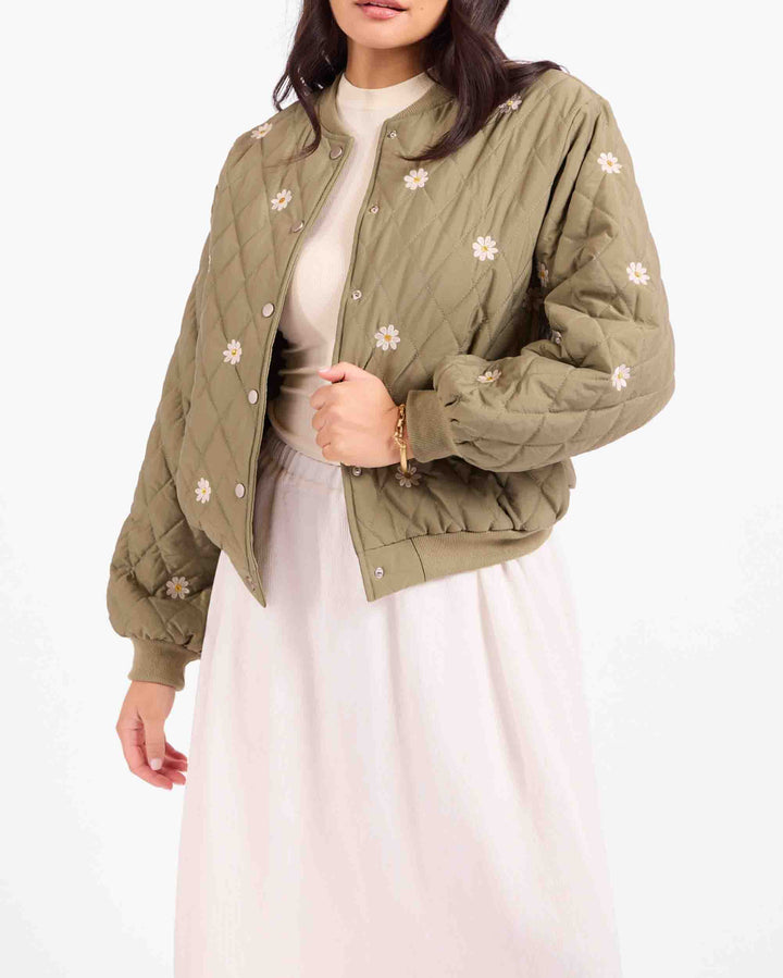 A woman wearing an olive green quilted bomber jacket with white embroidered flowers, styled over a cream top and a light pink skirt against a plain white background.