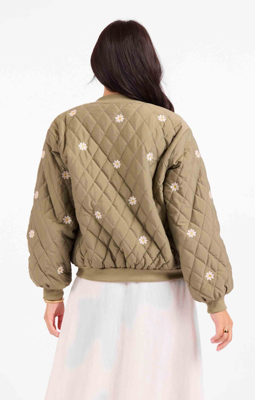 A woman viewed from the back wearing a quilted olive green bomber jacket with white embroidered floral details, paired with a light-colored skirt, against a plain white background.