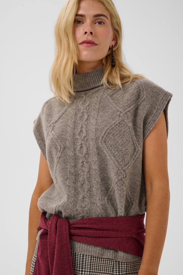 A woman wearing a gray cable-knit sleeveless mock-neck sweater, styled with a dark red tied waist sash and a black-and-white checkered skirt, against a light gray studio background.