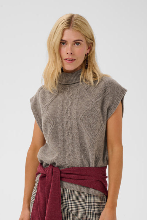 A blonde model wearing a gray cable-knit sleeveless turtleneck sweater vest, styled with a dark red sweater tied around her waist and a gray and black plaid skirt, against a light gray studio background.