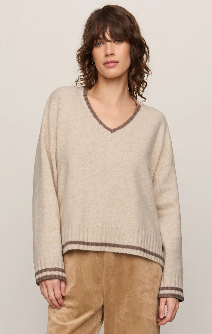 A woman wearing a light beige knit sweater with brown trim on the V-neck, cuffs, and hem, paired with tan pants, against a plain light beige background.