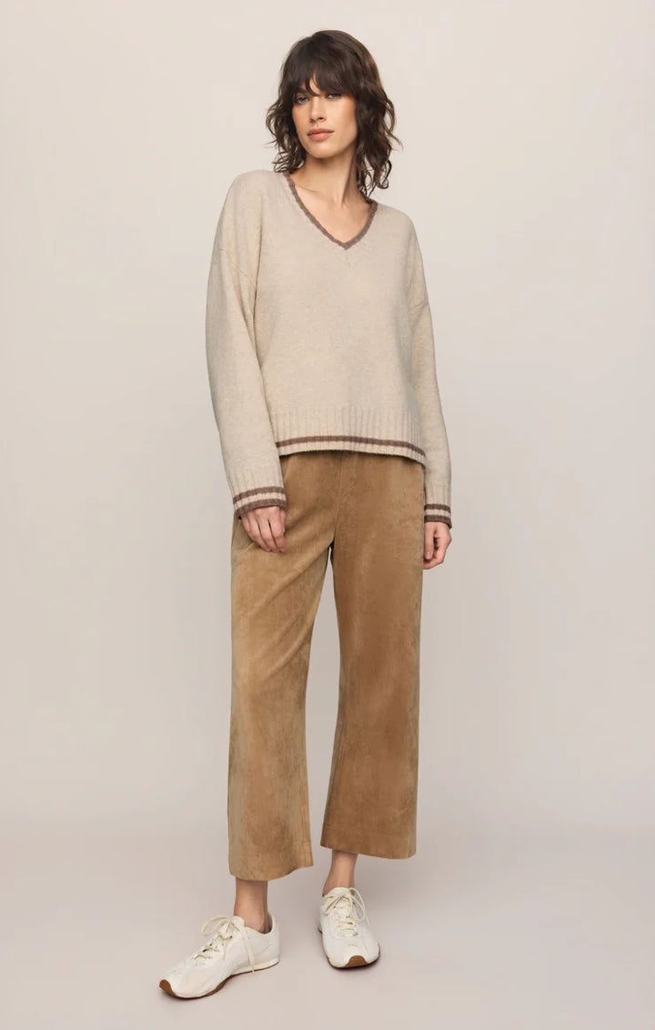 A model stands against a plain light beige studio background wearing a cream-colored v-neck knit sweater with brown striped trim, brown wide-leg cropped pants, and white sneakers.