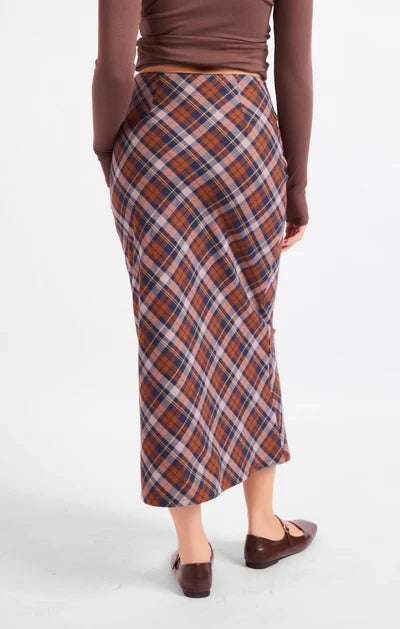 A woman shown from the back wearing a brown long-sleeve top and a rust-orange plaid midi skirt with navy and white lines, paired with brown flats, against a light gray studio background.