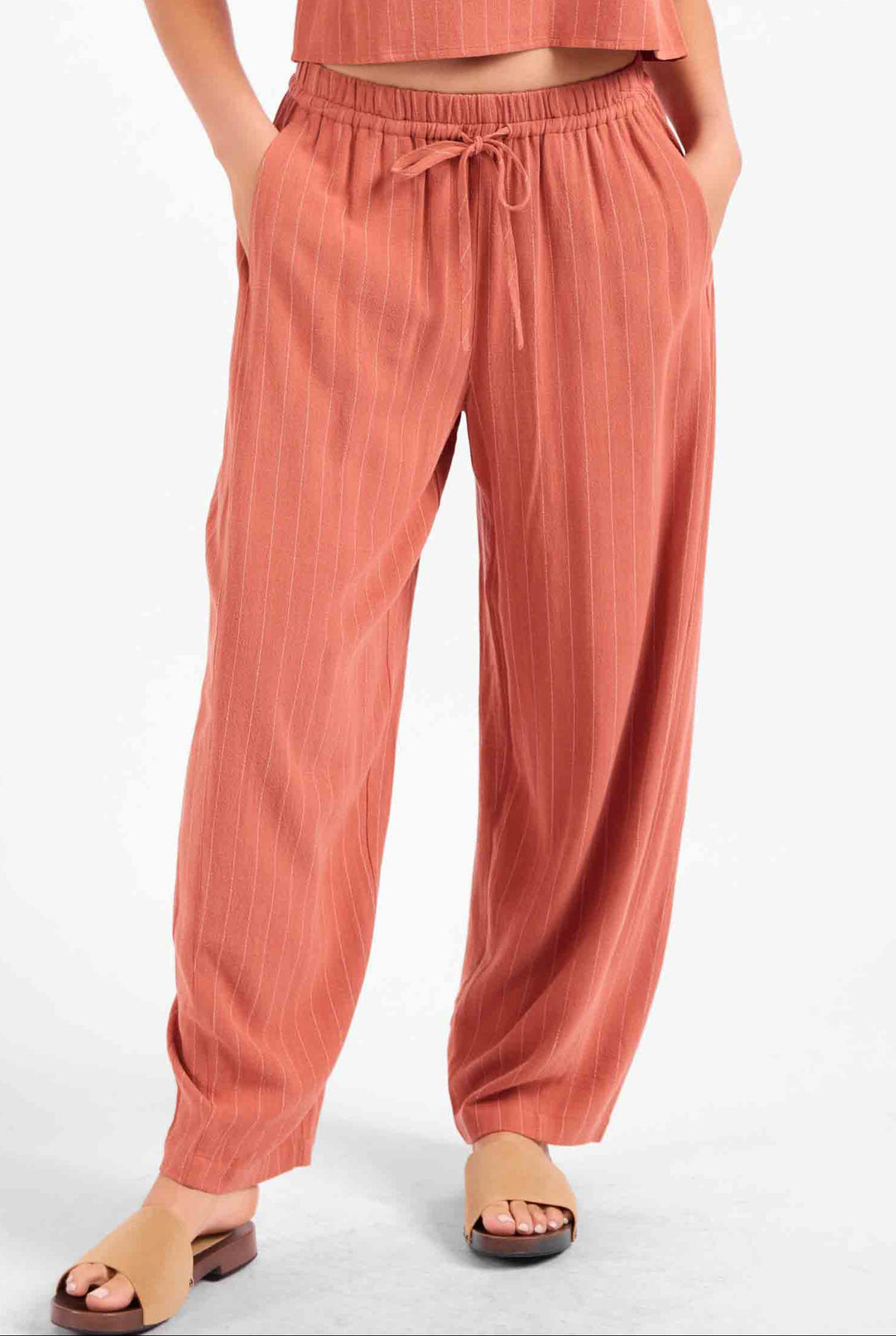 A person wearing loose, wide-leg drawstring pants in a muted terracotta or dusty rose tone, paired with a matching cropped top and tan slide sandals, against a plain light background.