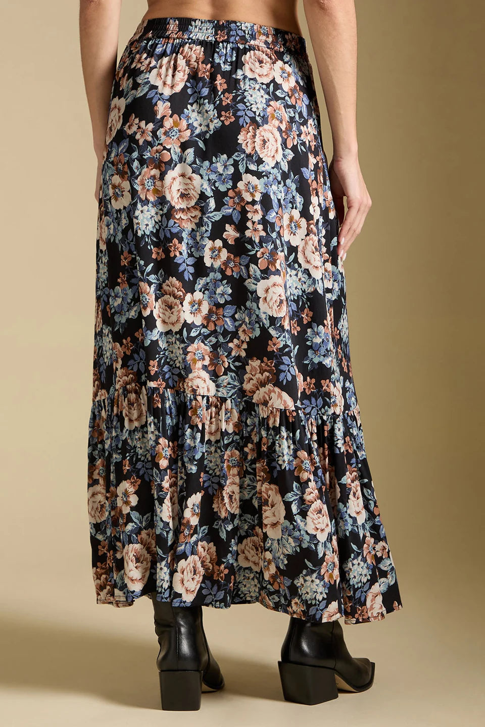 A model from the back wearing a black pleated maxi skirt with a large floral print in beige, pink, and blue, paired with black block-heel boots against a solid beige background.