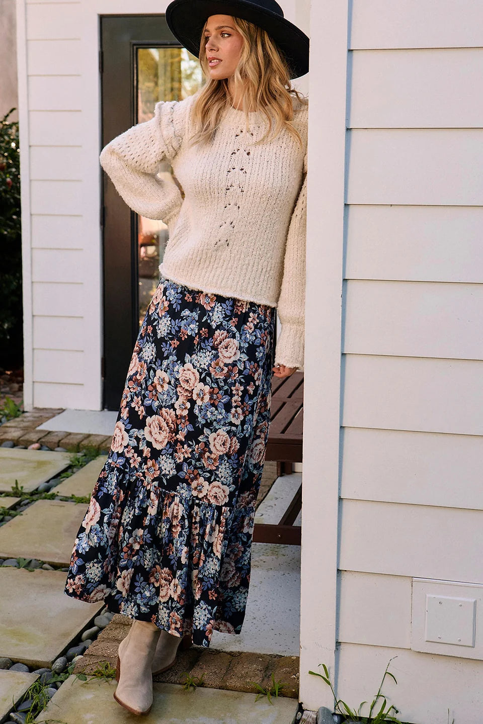 Woman in a floral skirt and beige sweater standing outside a house.